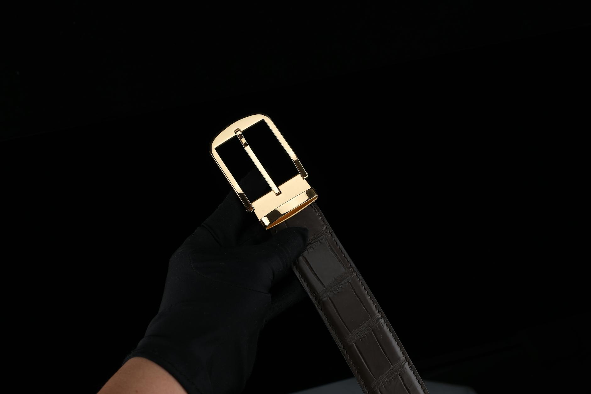 10 Accessory Mistakes Secretly Ruining Your Outfits (+ Fix) A close-up of a hand in glove holding a luxury leather belt with a golden buckle.